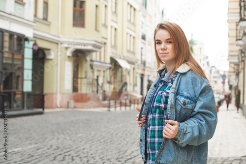 Beautiful lady in casual clothing is standing with a backpack in the background of a street landscape, looking into the camera and smiling. Street portrait of a beautiful stylish girl. Street fashion