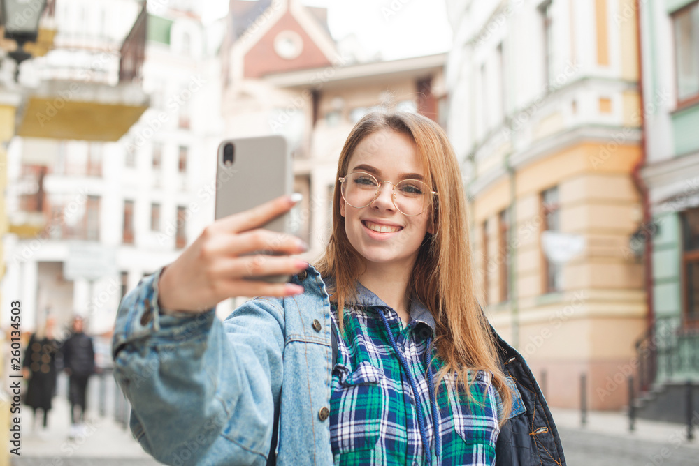 Happy girl traveler in casual clothing makes a selfie on the streets town, smiles and looks at the camera of a smartphone. Girl student pose on a smartphone camera, wears a jeans jacket and glasses.