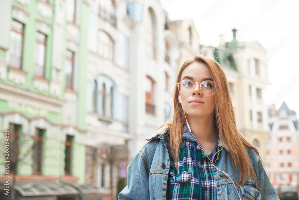 Portrait of a beautiful girl in casual clothing stands in the street with a serious face, listens to music in the headphones and looks side by side with the landscape of the town. Portrait of a beautiful girl in casual clothing stands in the street with a serious face, listens to music in the headphones and looks side by side with the landscape of the town.