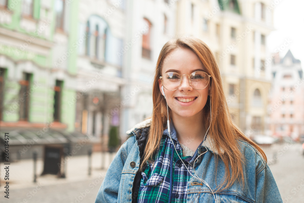 Portrait of a cheerful girl wearing casual clothing, standing in headphones, background of the landscape of the old town,looking camera and smiling.Close portrait of a call girl is listening to music