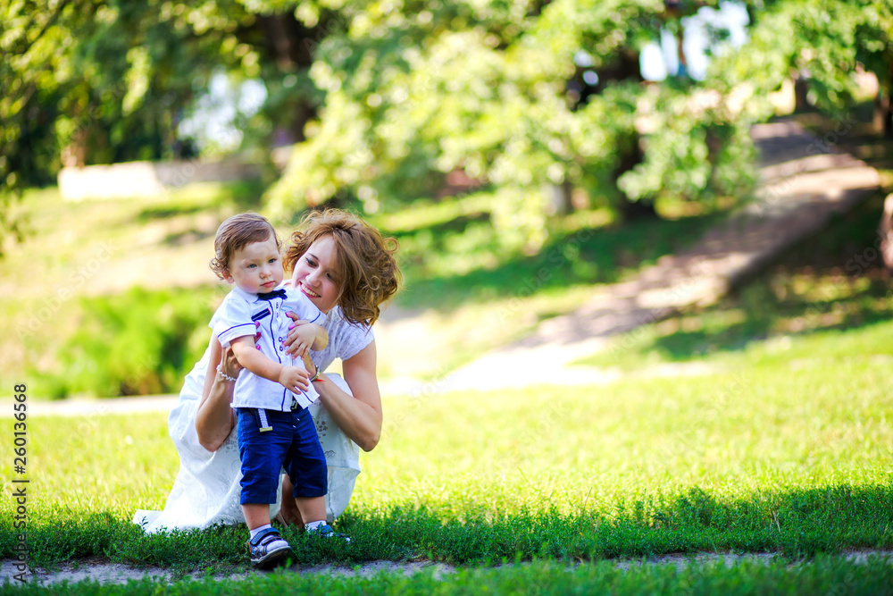 Fototapeta premium Mom hugs little son and playing in park at sunset time. People having fun on forest. Concept of friendly family and of summer vacation.