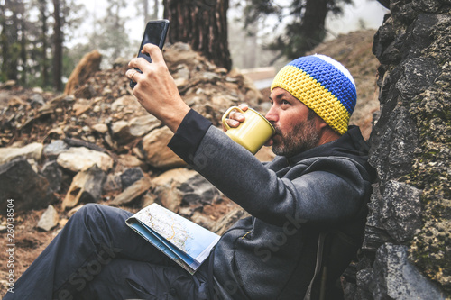 Bearded middle-aged man with woolen hood and sunglasses resting sitting after trekking day Male drinking coffee shooting selfie, chatting with friends. Relaxing with hot beverage in a National Park photo