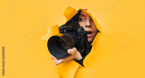 A young paparazzi girl with a black reflex camera looks out from cover and looks at what is happening with her mouth open. Yellow paper, torn hole. Tabloid press. In search of the plot for  stocks.