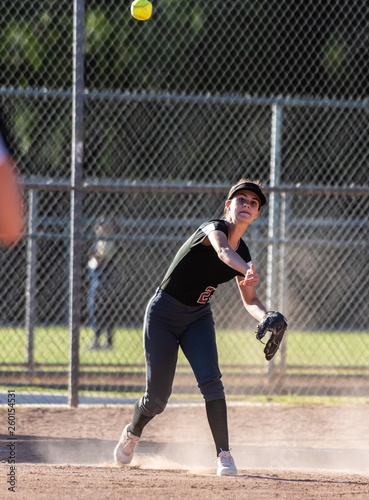 Female teenage softball player in black uniform throwing ball across infield for the out in a cloud of dust.