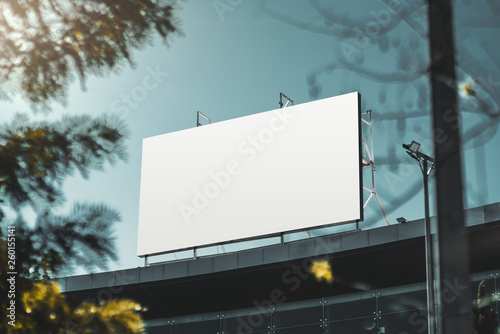 An empty huge poster mockup on the roof of a mall; white template placeholder of an advertising billboard on the rooftop of a modern building framed by trees; blank mock-up of an outdoor info banner photo