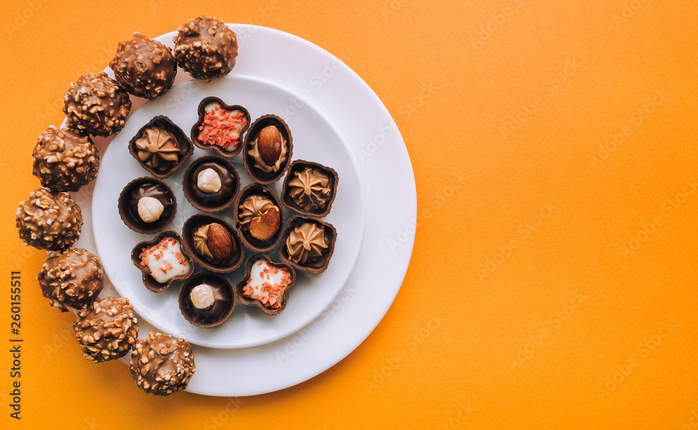 Beautiful chocolates of different shapes and fillings lie in a white plate against the background of orange paper. Top view, copy space. Beautiful chocolates of different shapes and fillings lie in a white plate against the background of orange paper. Top view, copy space.