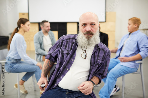 Portrait of bearded senior man looking at camera while sitting in chair in front of therapy group  copy space