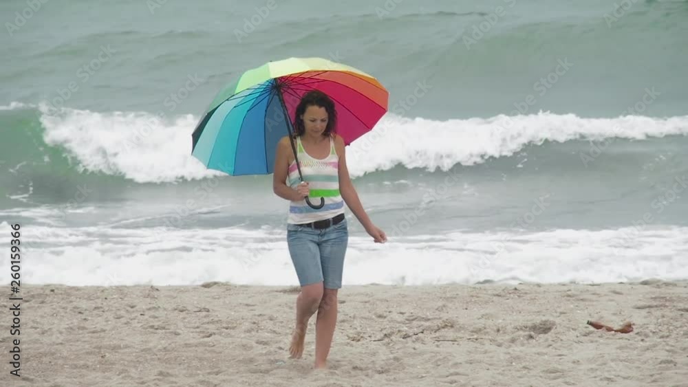 Woman walking in the rain on the beach. A female with a multi-colored umbrella walks along an empty beach. Woman walking in the rain on the beach. A female with a multi-colored umbrella walks along an empty beach.