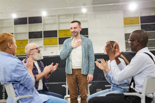 Portrait of smiling mature man    introducing himself during therapy session in support group to people clapping, copy space photo