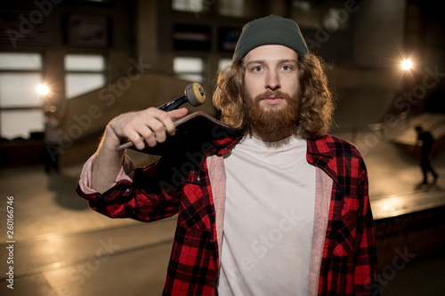 Waist up portrait of long haired man holding skateboard and looking at camera, shot with flash