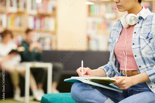 Close-up of serious busy student girl with headphones on neck making notes in workbook while doing homework in library