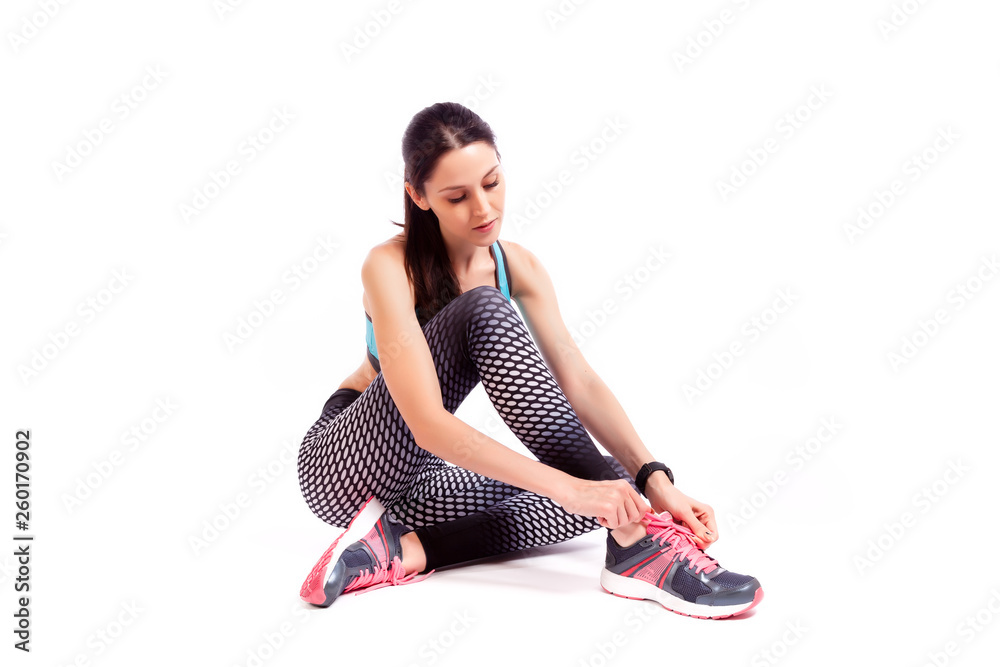 A young woman model in a sporty short top and gym leggings sitting on the floor and tying shoelaces on a white isolated background in studio A young woman model in a sporty short top and gym leggings sitting on the floor and tying shoelaces on a white isolated background in studio