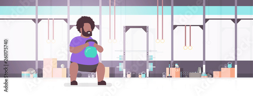 fat obese man doing exercises with kettlebell overweight fatty african american guy training workout weight loss concept modern gym health club studio interior horizontal full length flat