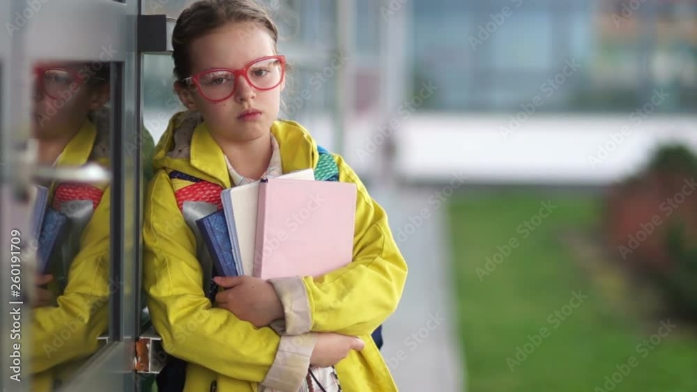 Close portrait of a tired schoolgirl with books in her hands. A girl in a yellow jacket, glasses with a red rim and a knapsack on her back is standing near the entrance to the school Close portrait of a tired schoolgirl with books in her hands. A girl in a yellow jacket, glasses with a red rim and a knapsack on her back is standing near the entrance to the school