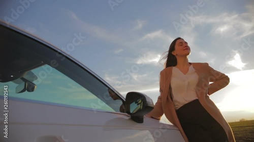 fashionable young woman stands near white car holding hand on hood against green meadow slow motion