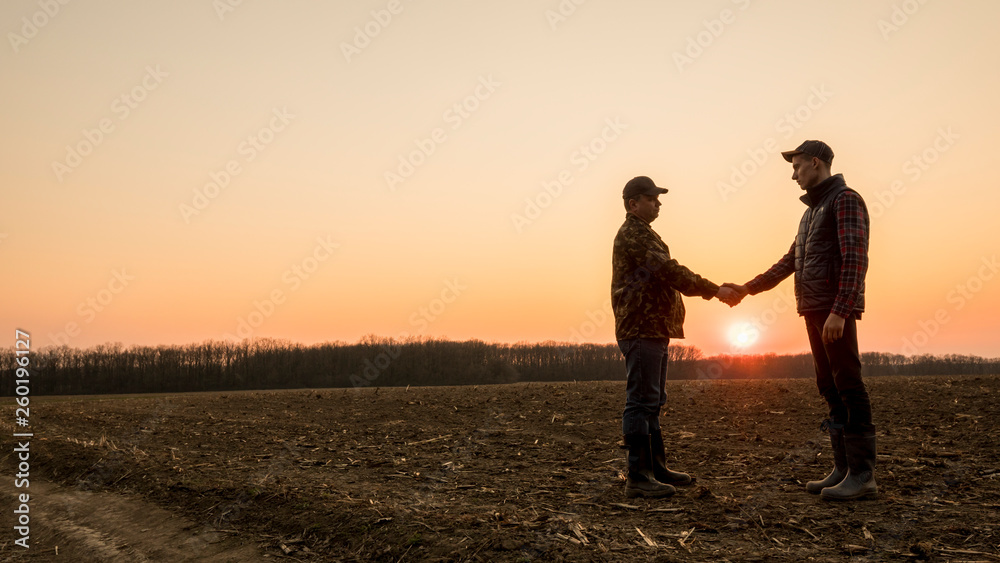 Two farmers on the field shake hands at sunset. Wide lens shot