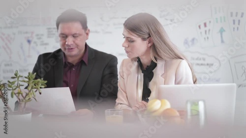 Static shot portrait of two young attractive business people sitting by the office desk, working together while focused on the paperwork, consulting an off screen coworker passing in the foreplan. photo