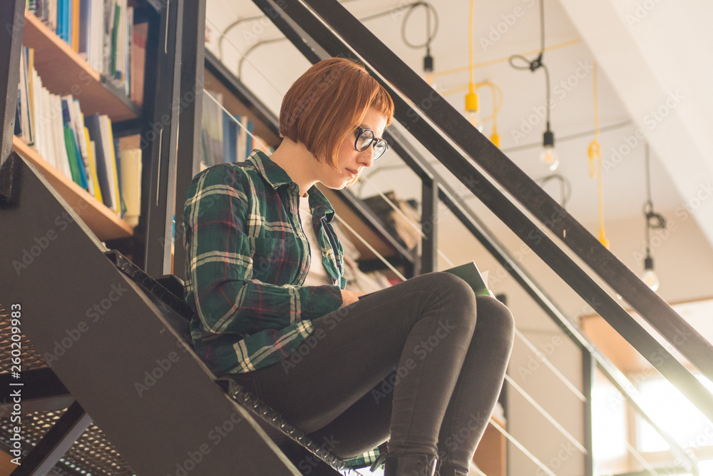 Young redhead woman in glasses read book in the library