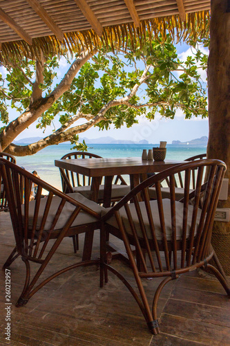 Empty table and chairs under trees on tropical beach. Beach cafe on seascape background. Outdoor restaurant wood furniture. Summer vacation concept. Travel in Asia. Idyllic dinner concept. Calm place.