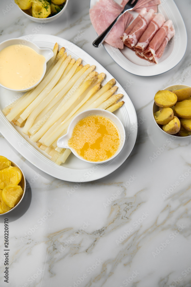 Platte weißer Spargel mit Sauce Hollandaise, zerlassener Butter, Kochschinken, geräucherten Schinken und Kartoffeln auf Marmor Hintergrund Platte weißer Spargel mit Sauce Hollandaise, zerlassener Butter, Kochschinken, geräucherten Schinken und Kartoffeln auf Marmor Hintergrund