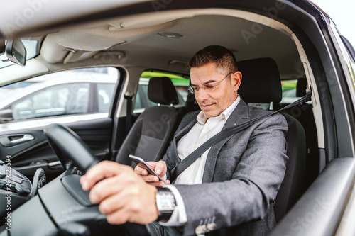 Smiling Caucasian man dressed smart casual with seat belt on and with hand on steering wheel suing smart phone for writing or reading message. Car interior.