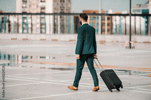 Ambitious Caucasian bearded businessman in formal wear carrying luggage and walking on parking lot. Business trip concept. Backs turned. Success occurs when your dreams get bigger than your excuses.