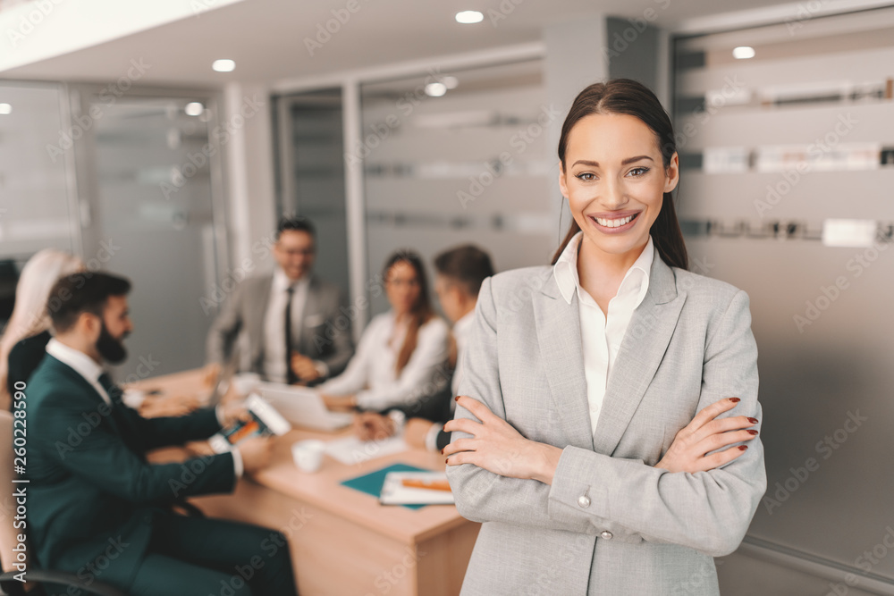 Beautiful Caucasian brunette in formal wear standing with arms crossed and smiling. In background colleagues having meeting. You don't build a business. Beautiful Caucasian brunette in formal wear standing with arms crossed and smiling. In background colleagues having meeting. You don't build a business.