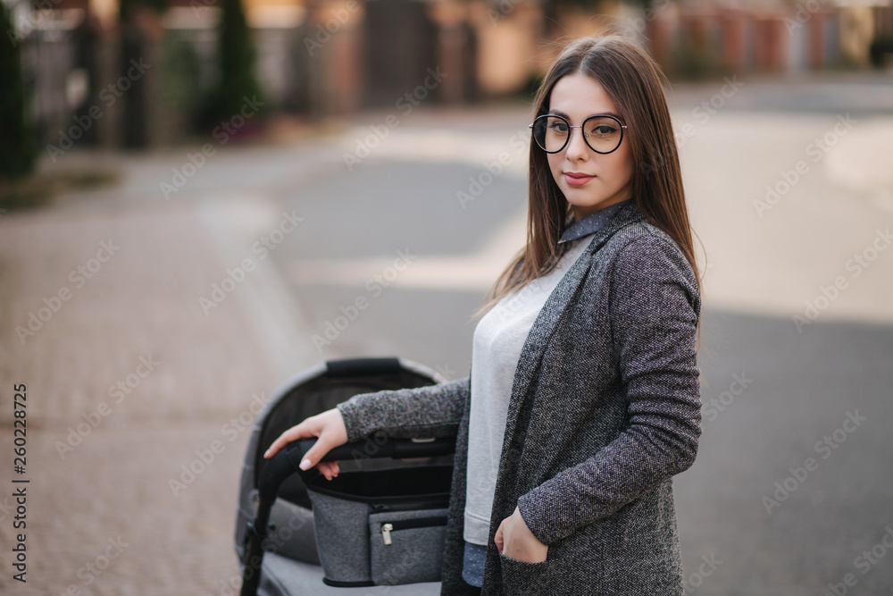 Family, child and parenthood concept - Happy mother with stroller in the park. Mom with sunglasses and stylish grey jacket Family, child and parenthood concept - Happy mother with stroller in the park. Mom with sunglasses and stylish grey jacket