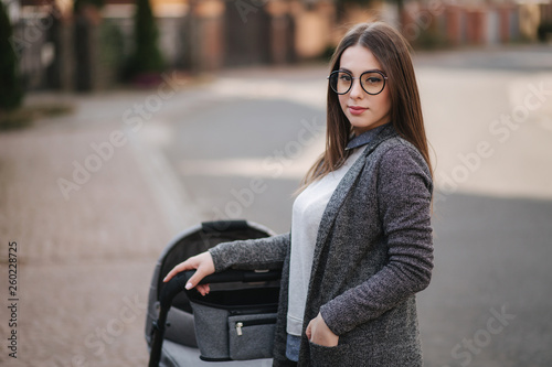 Family, child and parenthood concept - Happy mother with stroller in the park. Mom with sunglasses and stylish grey jacket © Aleksandr