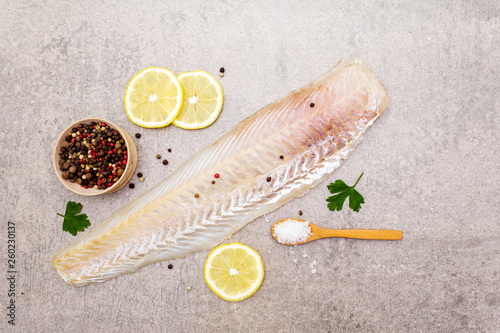 Fresh raw hake fillet on a stone background. With lemon, parsley, salt in wooden spoon and peppers mix. Top view