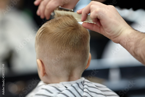 Cute blond baby boy in a barber shop having haircut by hairdresser. Hands of stylist with tools. Children's fashion. Indoors, back view, copy space.
