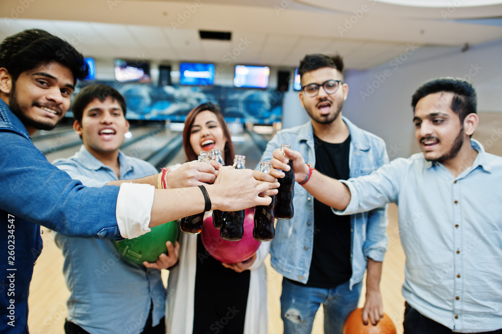 Obraz premium Group of five south asian peoples having rest and fun at bowling club. Clinking cold soda drinks from glass bottles and bowling balls at hands.