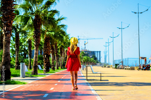 Young attractive woman in red dress walks along the pedestrian road with palm trees in the summer, near the Black Sea coast, Batumi, Georgia © volhavasilevich