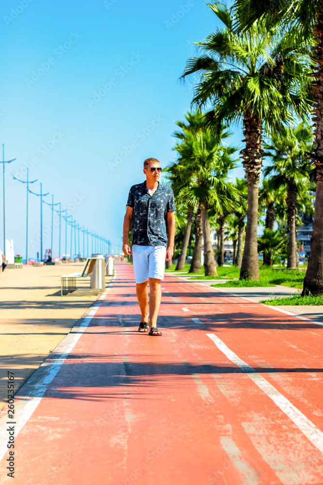 Young attractive guy walks along the pedestrian road with palm trees in the summer, near the Black Sea coast, Batumi, Georgia