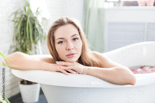 A young attractive girl takes a bathroom with flower petals and relaxes against the background of a beautiful light interior. Spa treatments for beauty and health with skin care