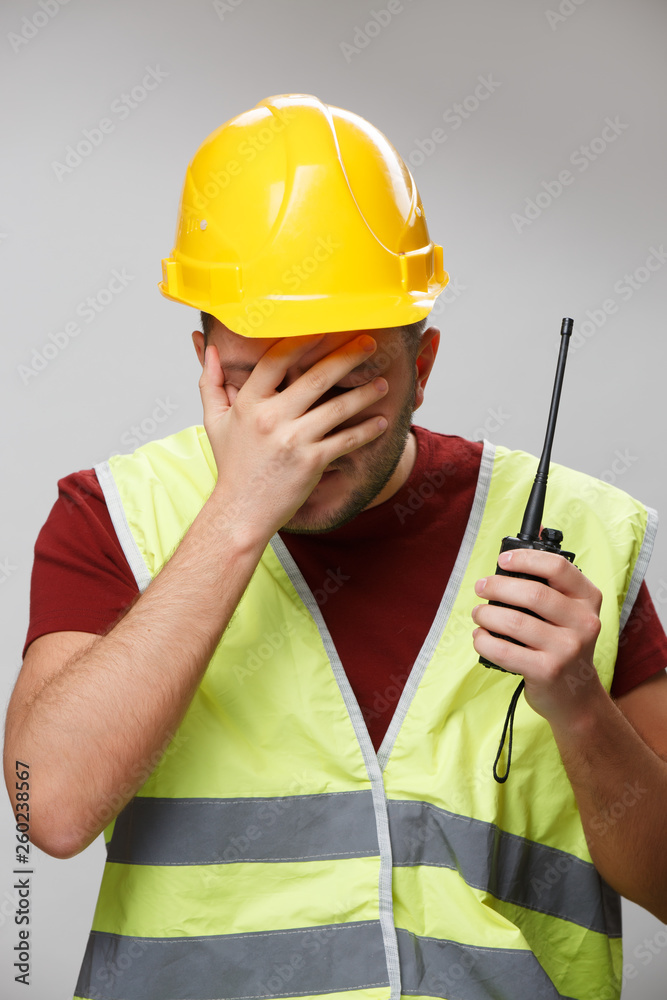 Photo of unhappy builder in yellow helmet with walkie-talkie on empty gray background.