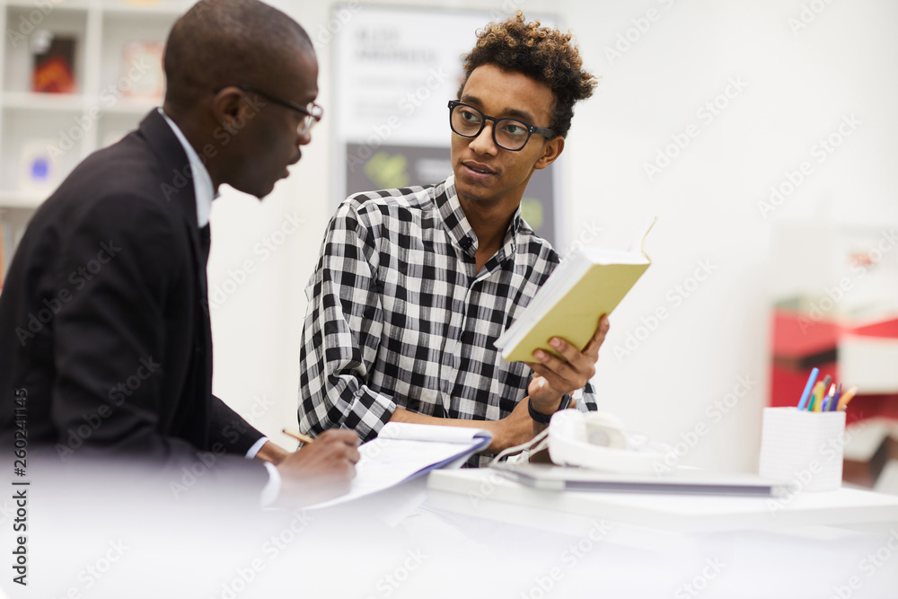 Serious pensive black guys in glasses sitting at table and discussing ideas for project: young man with Afro hairstyle reading aloud book while his groupmate making notes in paper