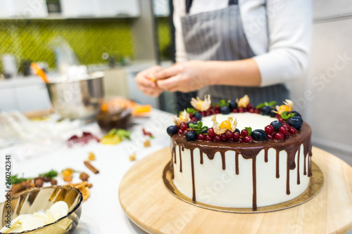 Confectioner decorates with berries a biscuit cake with white cream and chocolate. Cake stands on a wooden stand on a white table. The concept of homemade pastry  cooking cakes.