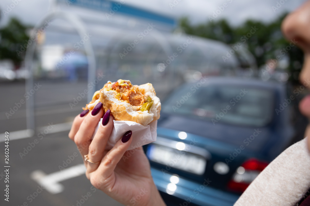A young woman holds a bitten hot dog in her hand. Snack in the parking lot near the shopping center.