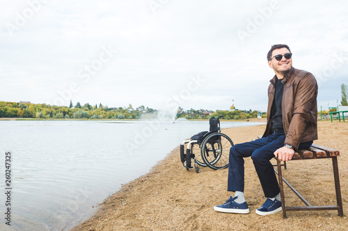 A young man sits on a bench by the lake, next to his wheelchair.