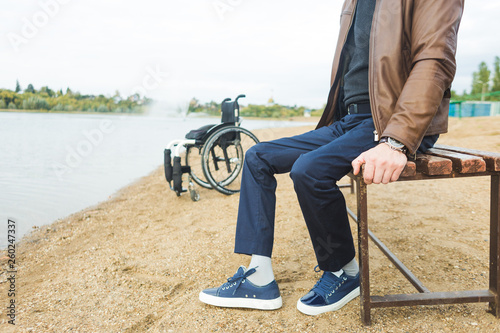 A young man sits on a bench by the lake, next to his wheelchair.