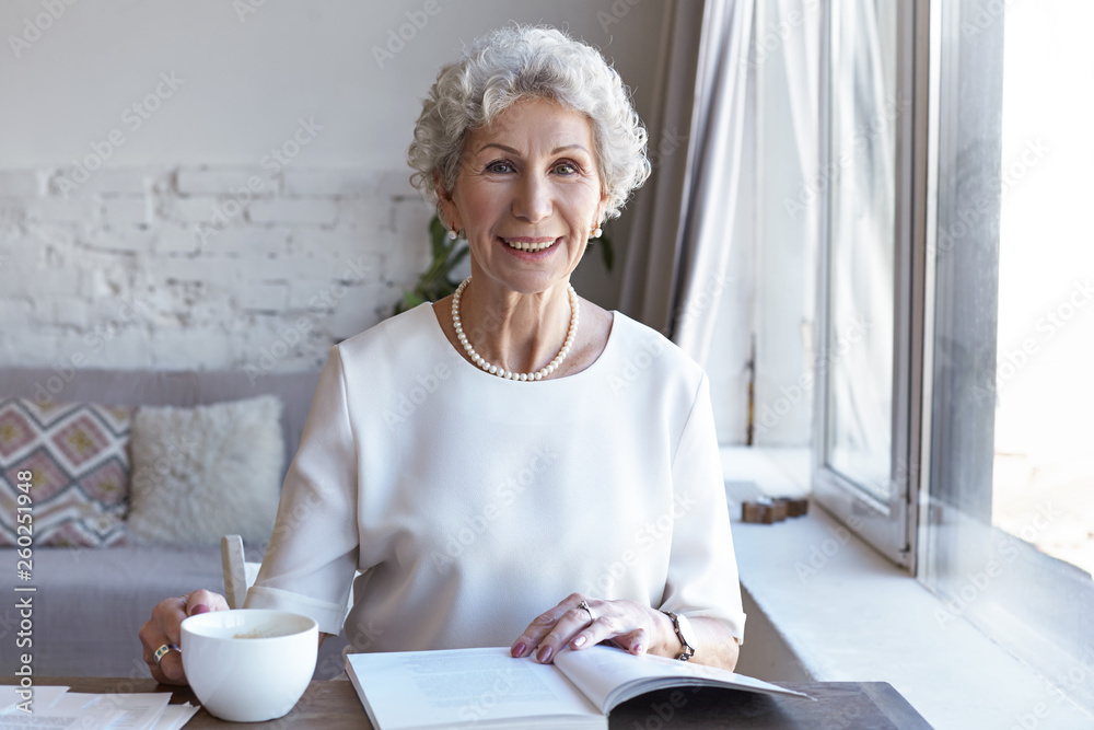Horizontal shot of cheerful happy retired European gray haired lady relaxing indoors with cup of fresh hot coffee and interesting book. Charming female pensioner reading article in fashion magazine Horizontal shot of cheerful happy retired European gray haired lady relaxing indoors with cup of fresh hot coffee and interesting book. Charming female pensioner reading article in fashion magazine