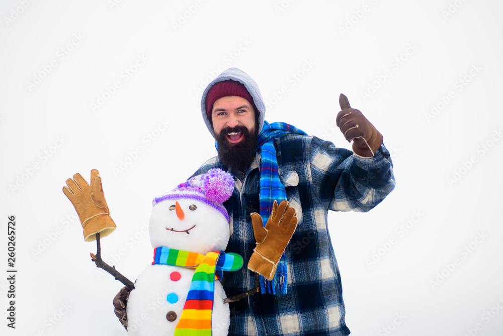 Winter man standing near snowman. Bearded man shows thumb up. New year snowman in knitted hat&striped scarf. Happy holiday&celebration. Man in winter clothes raised thumb up. Happy man hugs snowman. Winter man standing near snowman. Bearded man shows thumb up. New year snowman in knitted hat&striped scarf. Happy holiday&celebration. Man in winter clothes raised thumb up. Happy man hugs snowman.