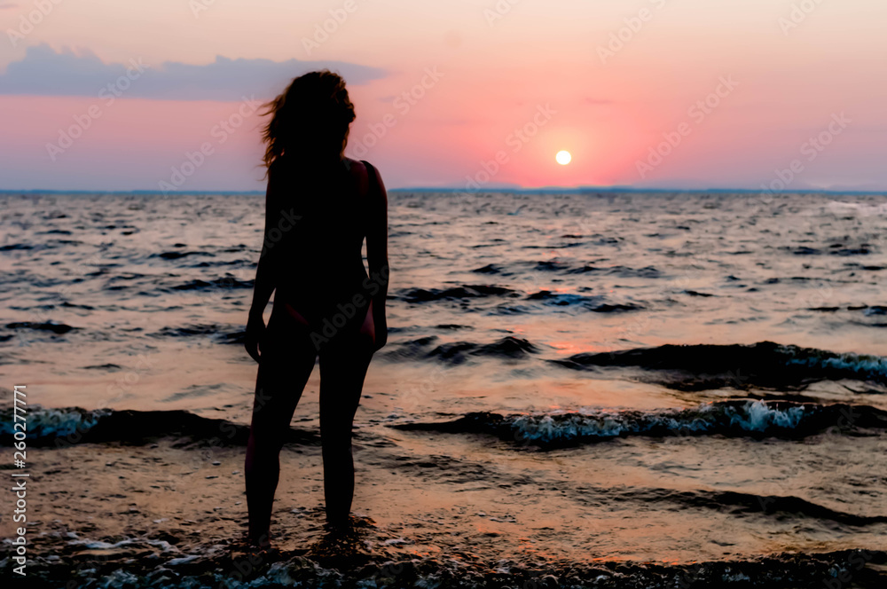 back view of slim woman figure in swimsuit standing and looking at amazing sunrise near sea on beach