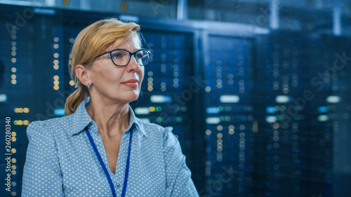 Portrait of a Beautiful Senior Female IT Technician Standing In Modern Data Center. In the Background Working Server Racks with Blinking LED Lights.
