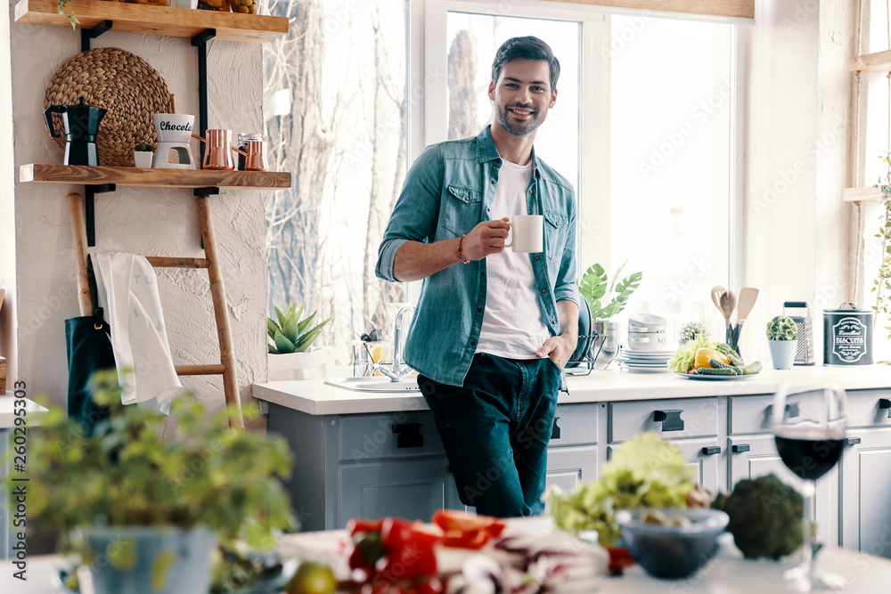 Modern man. Handsome young man in casual wear looking at camera and having a hot drink while standing in the kitchen at home Modern man. Handsome young man in casual wear looking at camera and having a hot drink while standing in the kitchen at home
