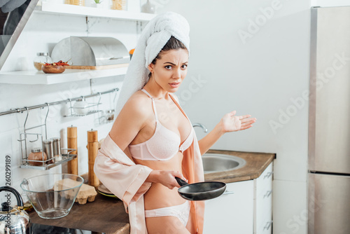 Dissatisfied sexy girl in housecoat with towel on head holding frying pan in kitchen