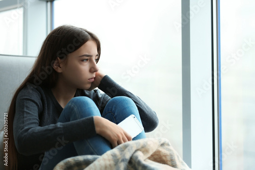 Upset teenage girl with smartphone sitting at window indoors. Space for text