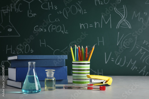 Laboratory glassware and school supplies on table against blackboard with chemical formulas