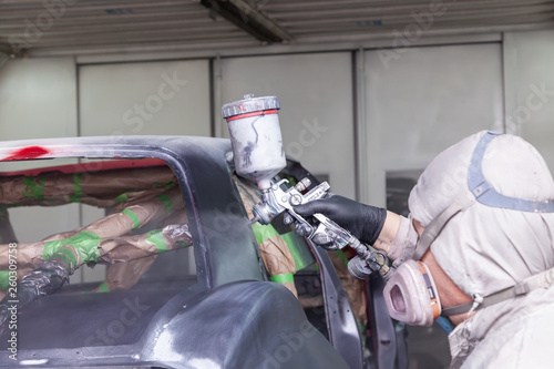 A man in protective overalls and a mask holds a spray bottle in his hand and sprays black paint onto the frame of the car body after an accident during a repair in a vehicle restoration workshop